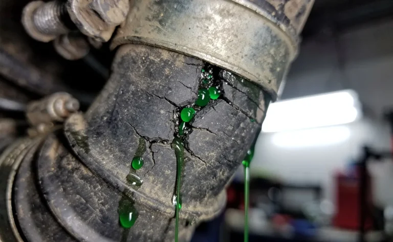 Gros plan sur un tuyau de climatisation usé d’une voiture, montrant des fissures et des fuites de réfrigérant vert sous l’éclairage d’un atelier de mécanicien.